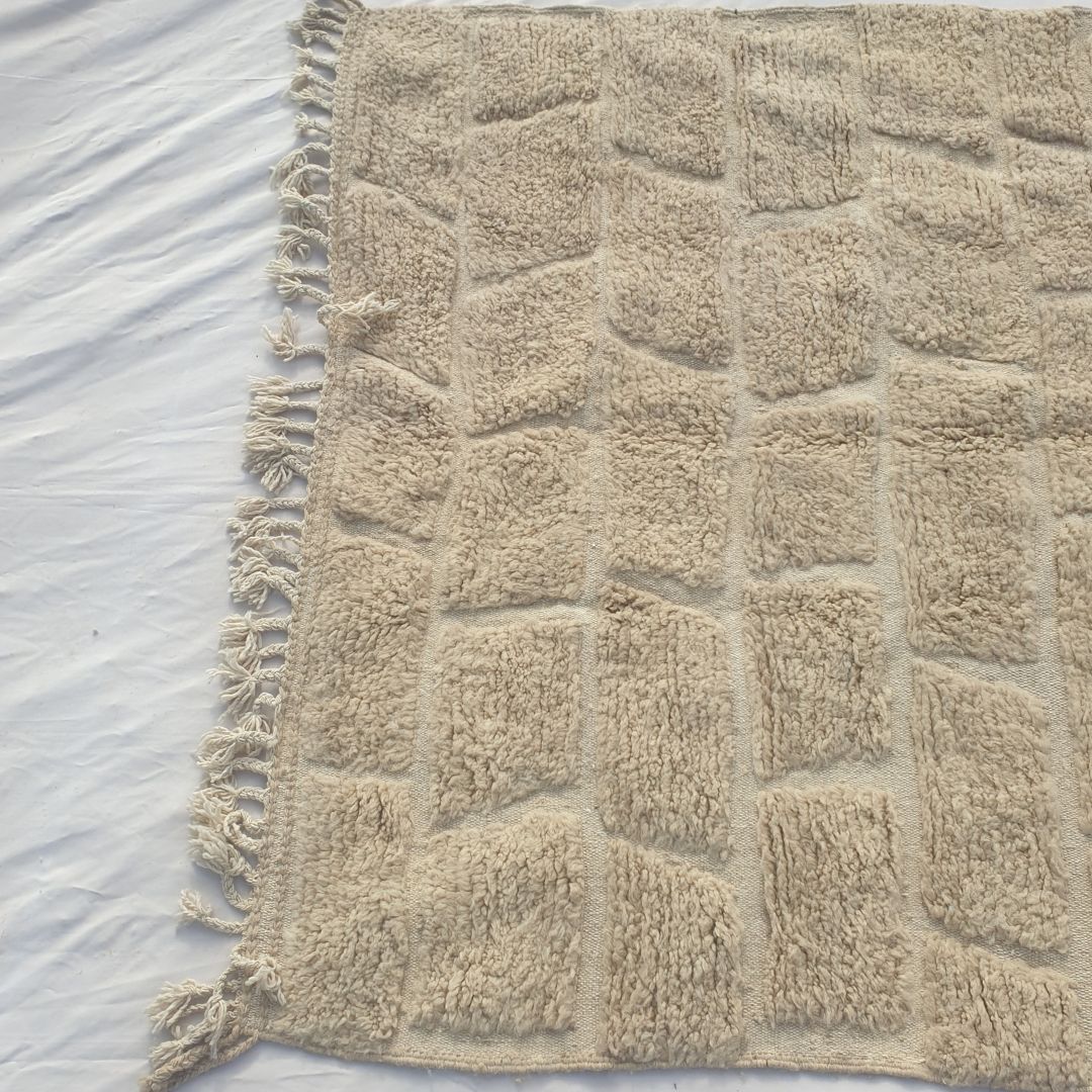 Detail shot of the ivory Beni Ourain brick cobblestone relief pattern on Yabre - the sculptural quality of the carved pile blocks visible in angled light, rows of rectangular forms with subtly textured surfaces separated by clean recessed lines, an architectural texture entirely in natural ivory wool