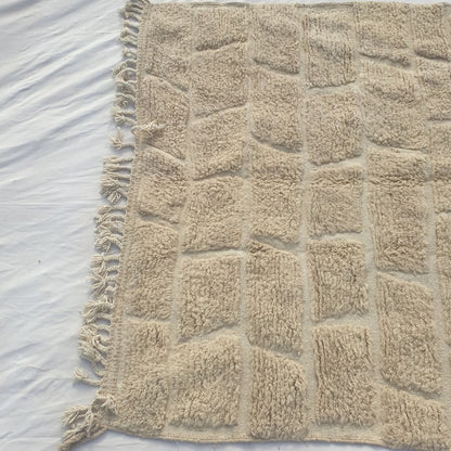 Detail shot of the ivory Beni Ourain brick cobblestone relief pattern on Yabre - the sculptural quality of the carved pile blocks visible in angled light, rows of rectangular forms with subtly textured surfaces separated by clean recessed lines, an architectural texture entirely in natural ivory wool