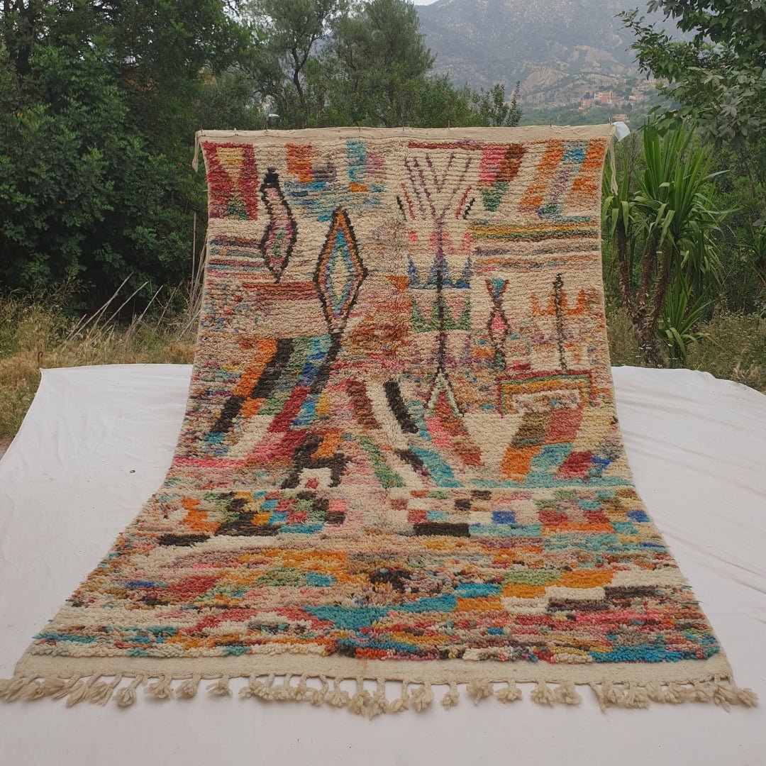Person holding up a colorful ivory Moroccan rug outdoors, displaying its 187x307 cm size - a dense ivory field covered in multicolor confetti-like flecks of turquoise, orange, pink and red, with a black-outlined diamond motif, a zigzag chevron pattern and a bold checkerboard band running through the middle